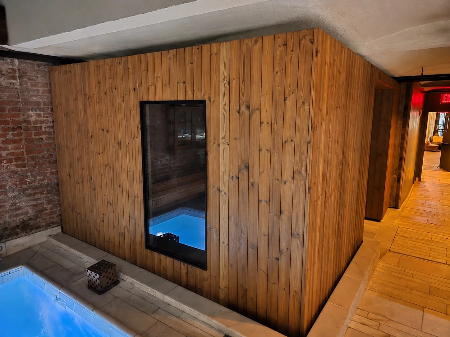AIRE Ancient Baths sauna from outside: a vertical timber-clad enclosure with a tall glass window beside a lit pool and a brick wall