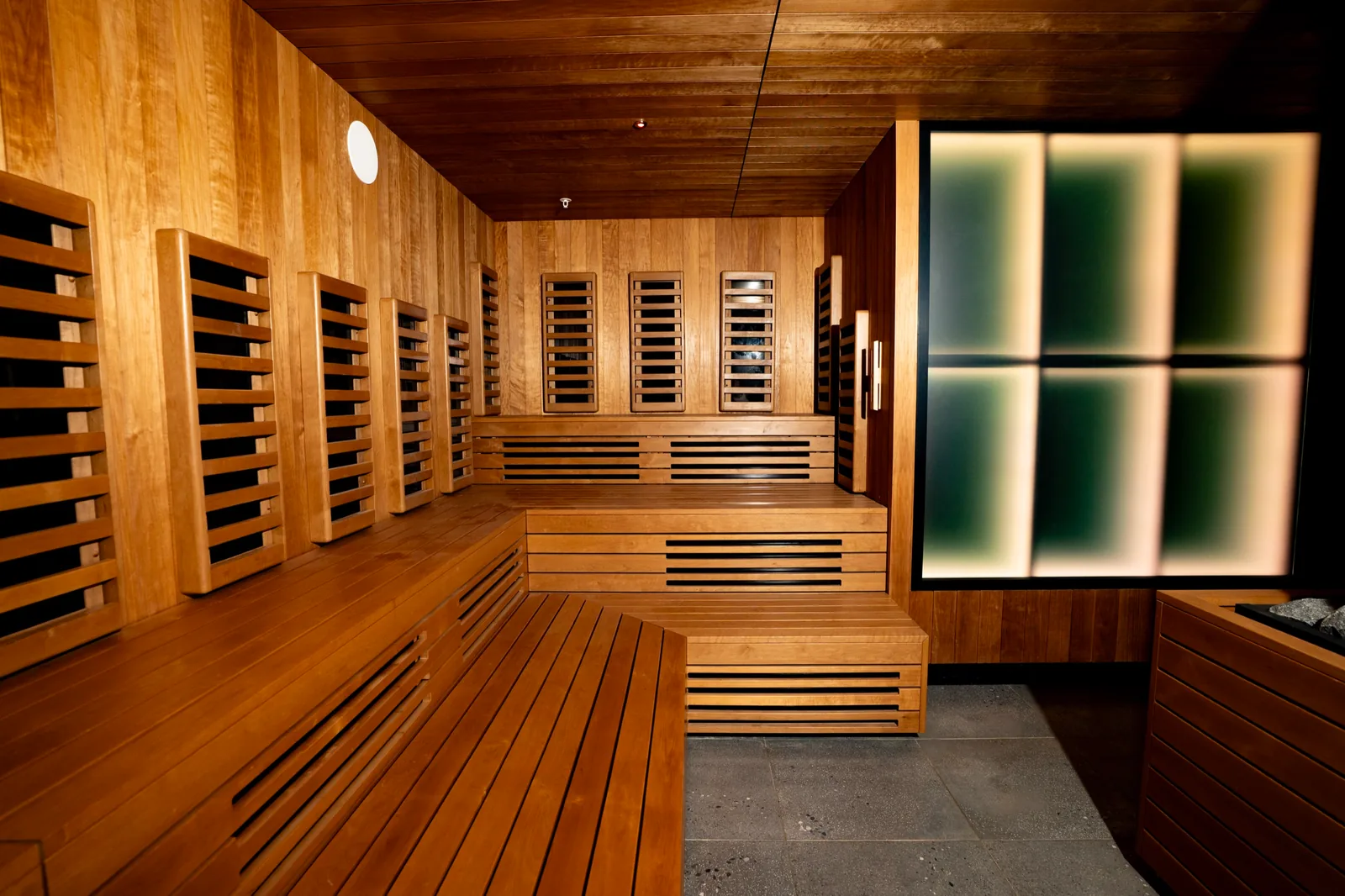 Bathhouse Atlantic infrared room: timber benches below infrared wall panels, with backlit frosted glass at the end of the room