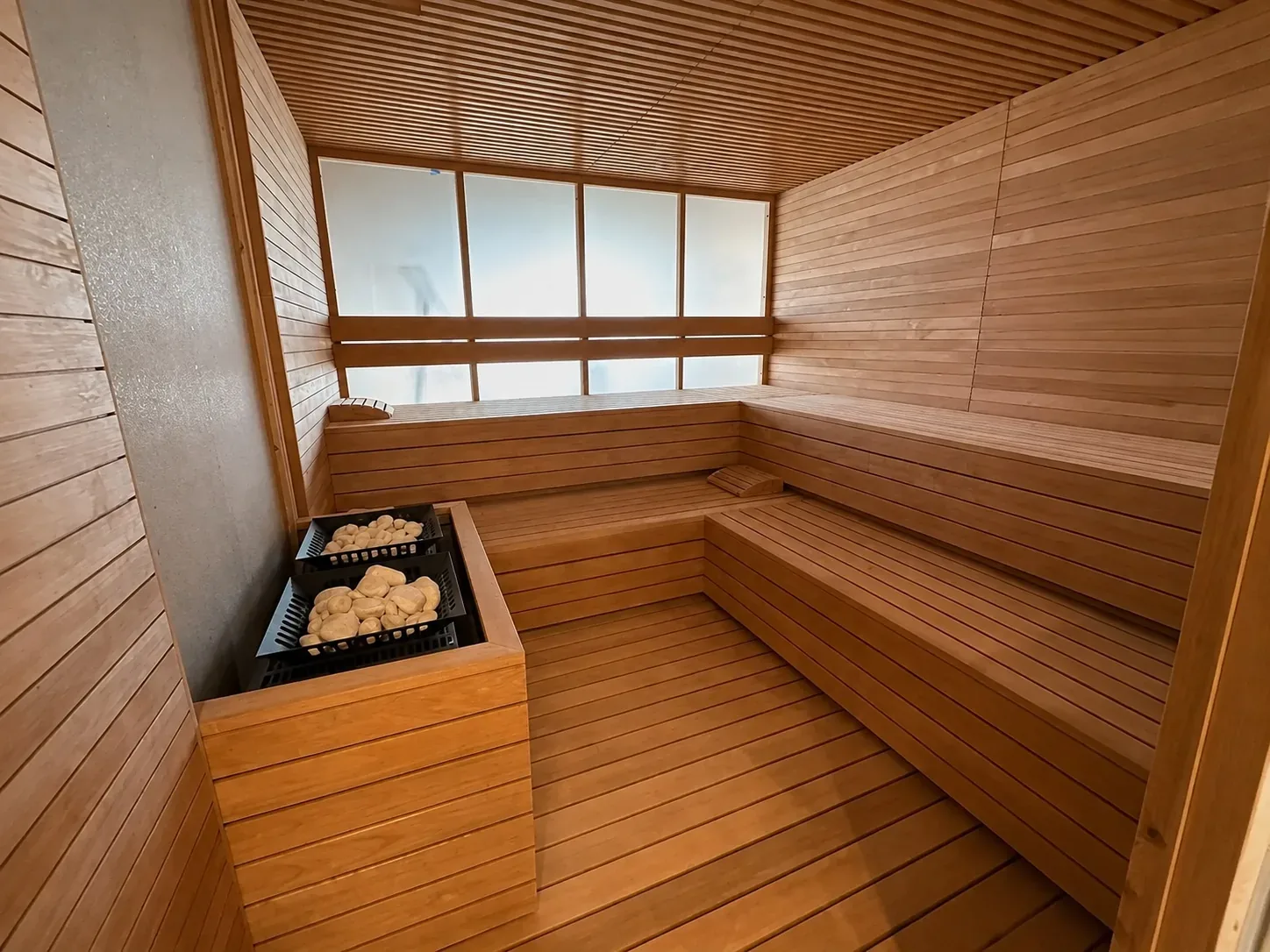 Cold Spring home sauna: L-shaped timber benches in two tiers, two stone-filled heater baskets in a timber surround and a frosted window wall